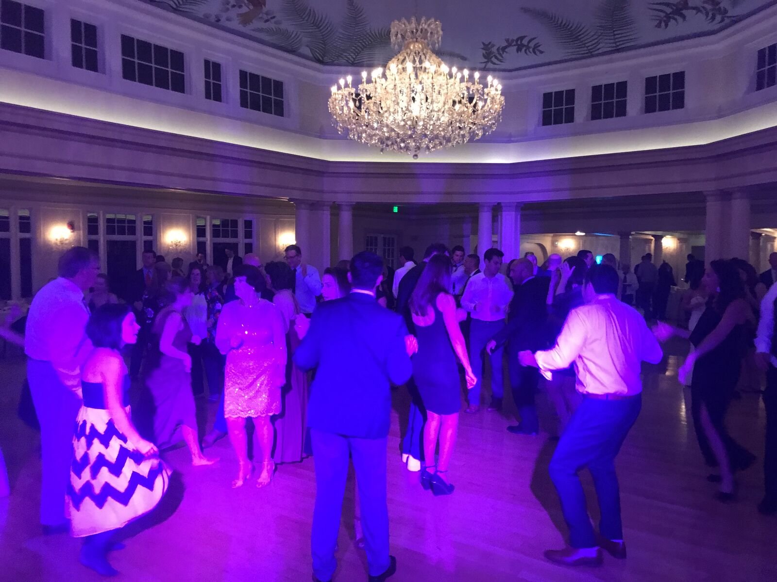Wedding guests dancing under a crystal chandelier in a ballroom lit with purple uplighting